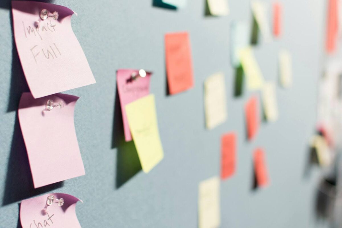 Colorful sticky notes are pinned to a bulletin board, some with handwritten text, arranged in a loose, unorganized pattern—capturing the creative energy of a design consultancy workspace.